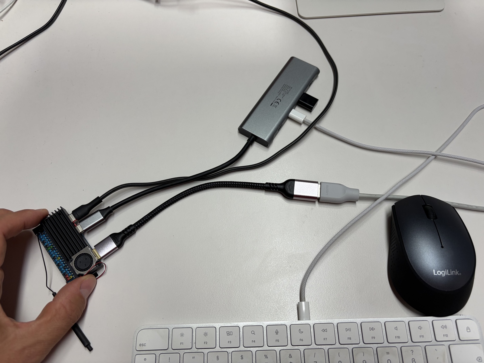 The Orange Pi Zero 3W held between two fingers on a desk, wearing a black finned heatsink and a small round fan over the SoC, with USB-C adapters for power and a hub plugged into both of its ports and a mini-HDMI cable running to a monitor - the whole board is shorter than the space bar of the Apple keyboard lying next to it