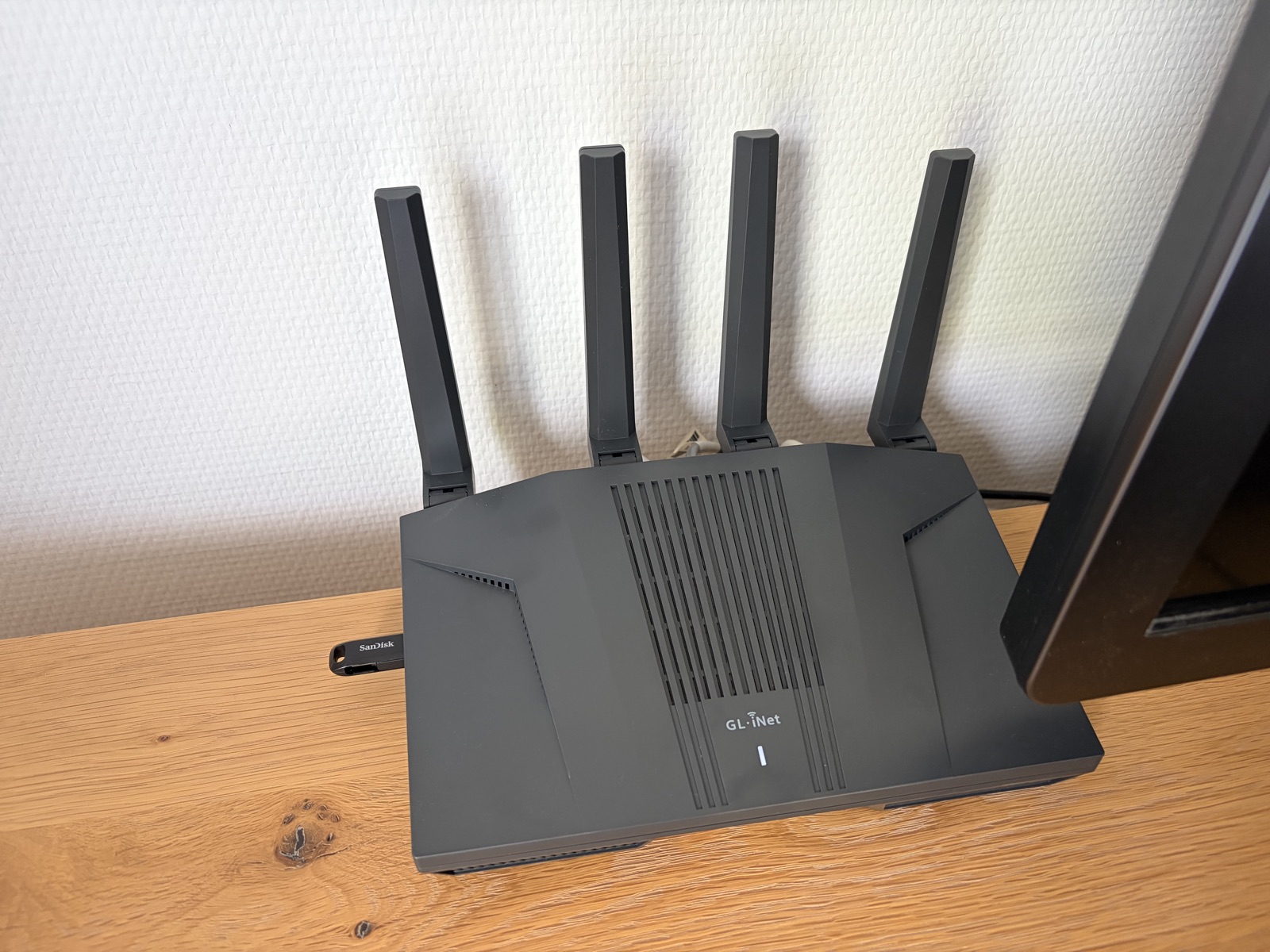 A GL.iNet Flint 2 GL-MT6000 router standing on a wooden desk against a white wall, its four black antennas raised, the GL.iNet logo and a single white status LED on the front face, and a SanDisk USB stick plugged into the port on its left side next to the foot of a monitor